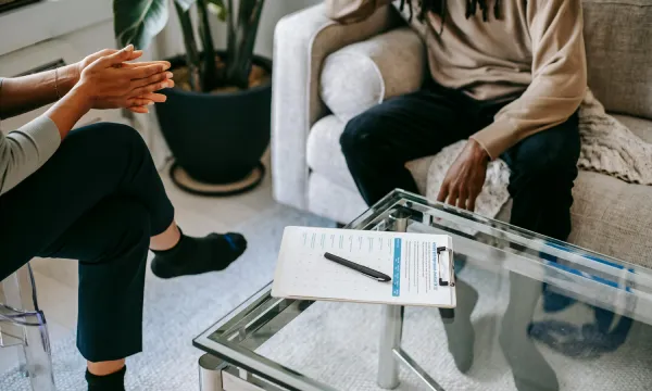 Two people in conversation seated near a glass coffee table, therapy notes visible